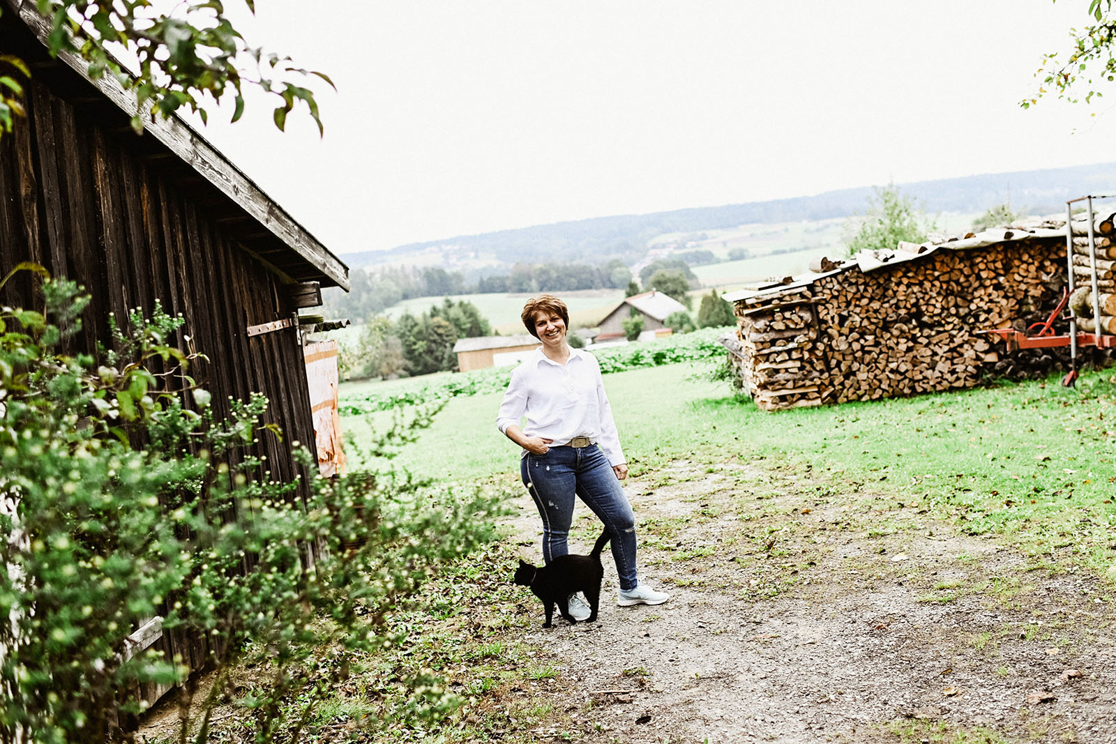 Naturheilpraxis Bianca Ahollinger, die Heilpraktikerin neben Holzschuppen im Garten mit schwarzer Katze